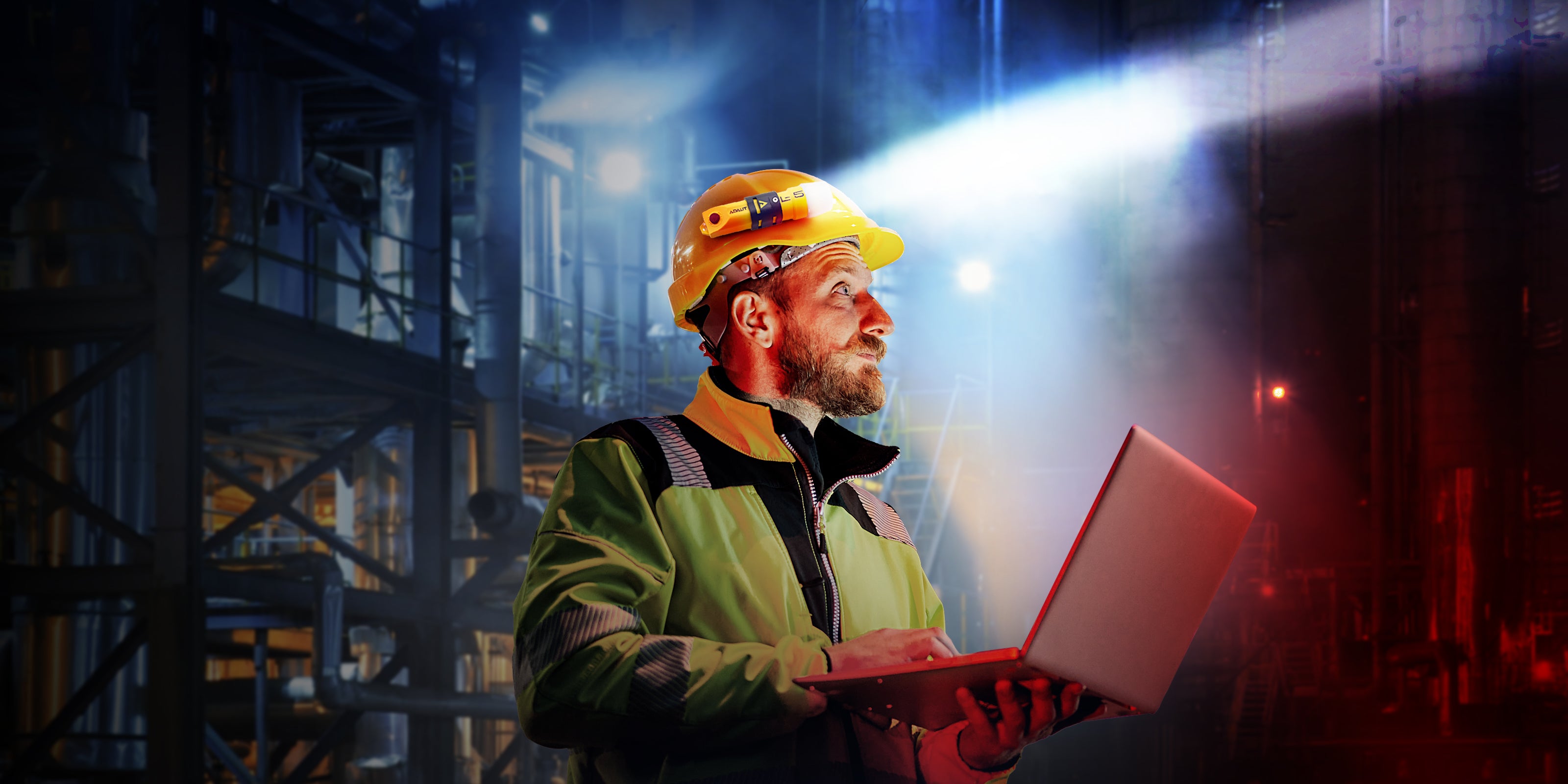 Person in a hard hat and safety jacket holding a laptop in an industrial setting